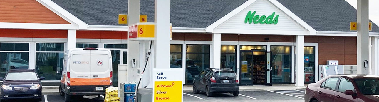 A photographed Needs store with cars visiting the gas pumps and customers entering the store.