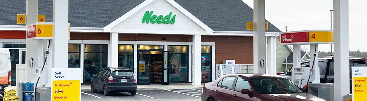 A photographed Needs store with cars visiting the gas pumps and customers entering the store.